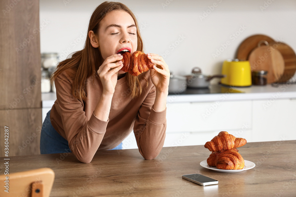 Beautiful young woman eating tasty croissant in kitchen