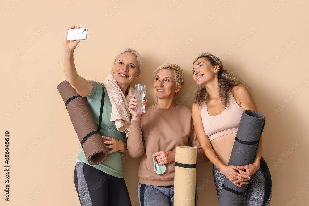 Sporty mature women with yoga mats taking selfie near beige wall