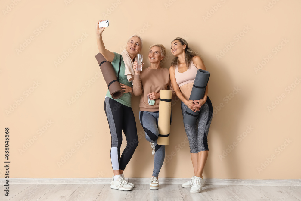 Sporty mature women with yoga mats taking selfie near beige wall