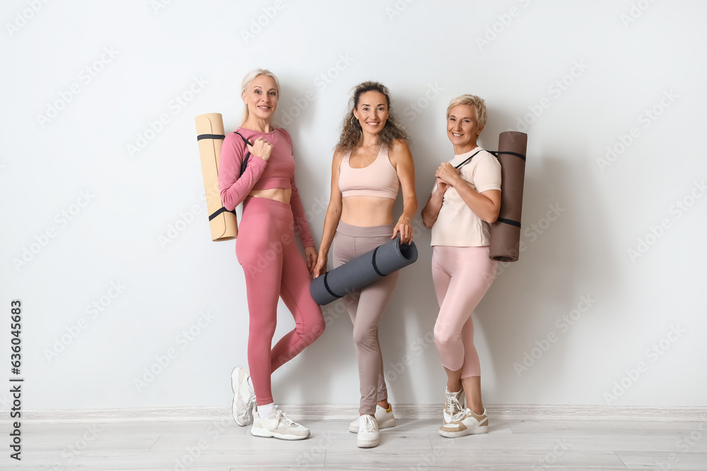 Sporty mature women with yoga mats near light wall