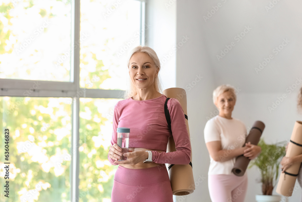 Sporty mature woman with yoga mat and bottle of water in gym