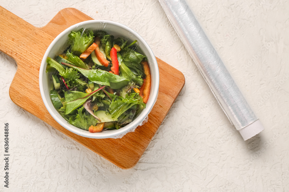 Bowl of fresh salad covered with plastic food wrap on light background