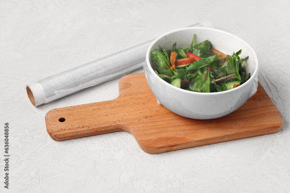 Bowl of fresh salad covered with plastic food wrap on light background