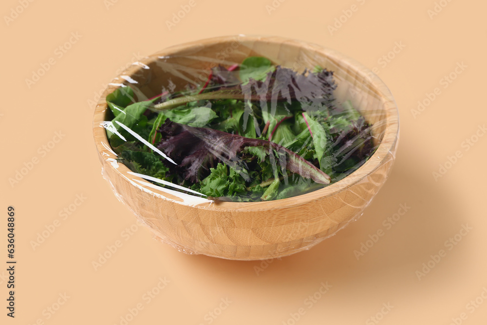 Bowl of fresh salad covered with plastic food wrap on beige background