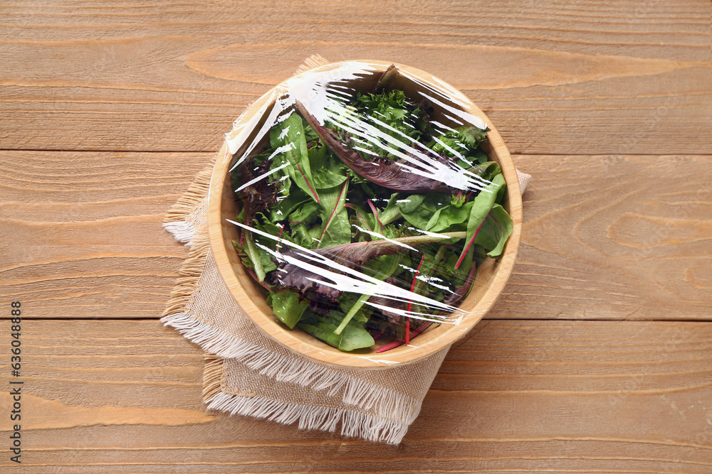 Bowl of fresh salad covered with plastic food wrap on wooden background