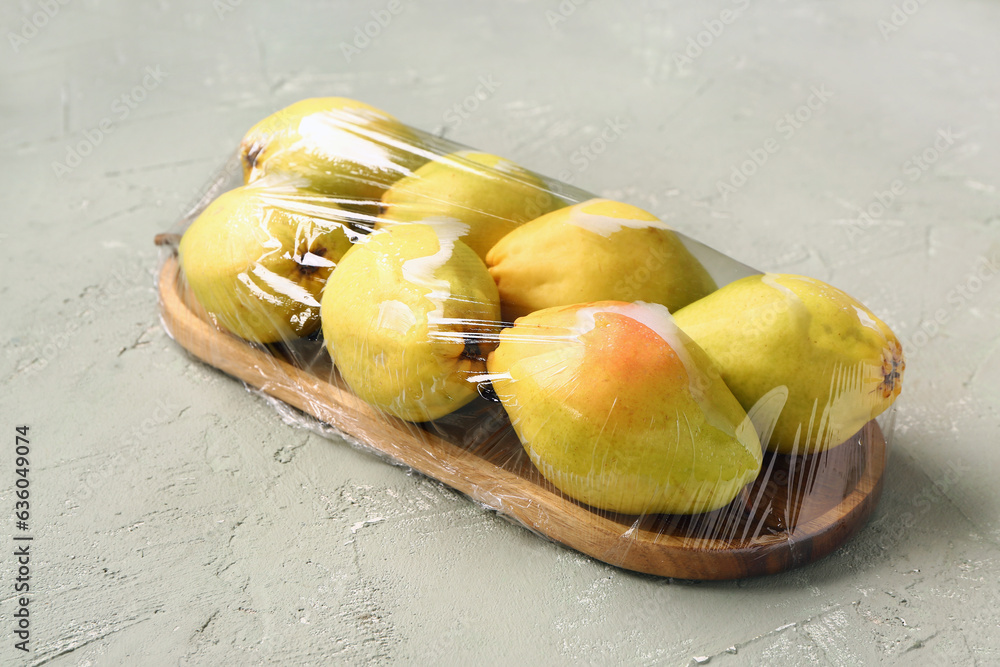 Wooden board of fresh pears covered with plastic food wrap on grey background