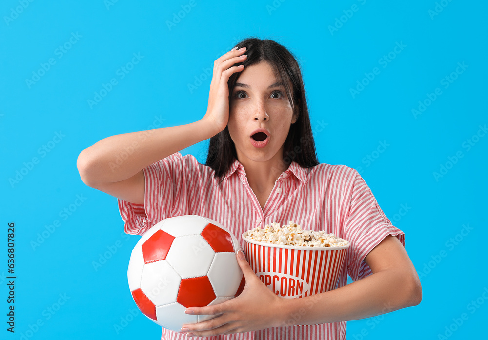 Shocked female soccer fan with popcorn and ball on blue background