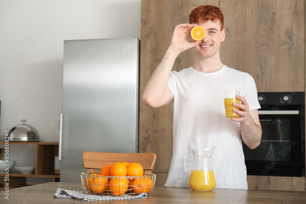 Young redhead man with orange and glass of juice in kitchen