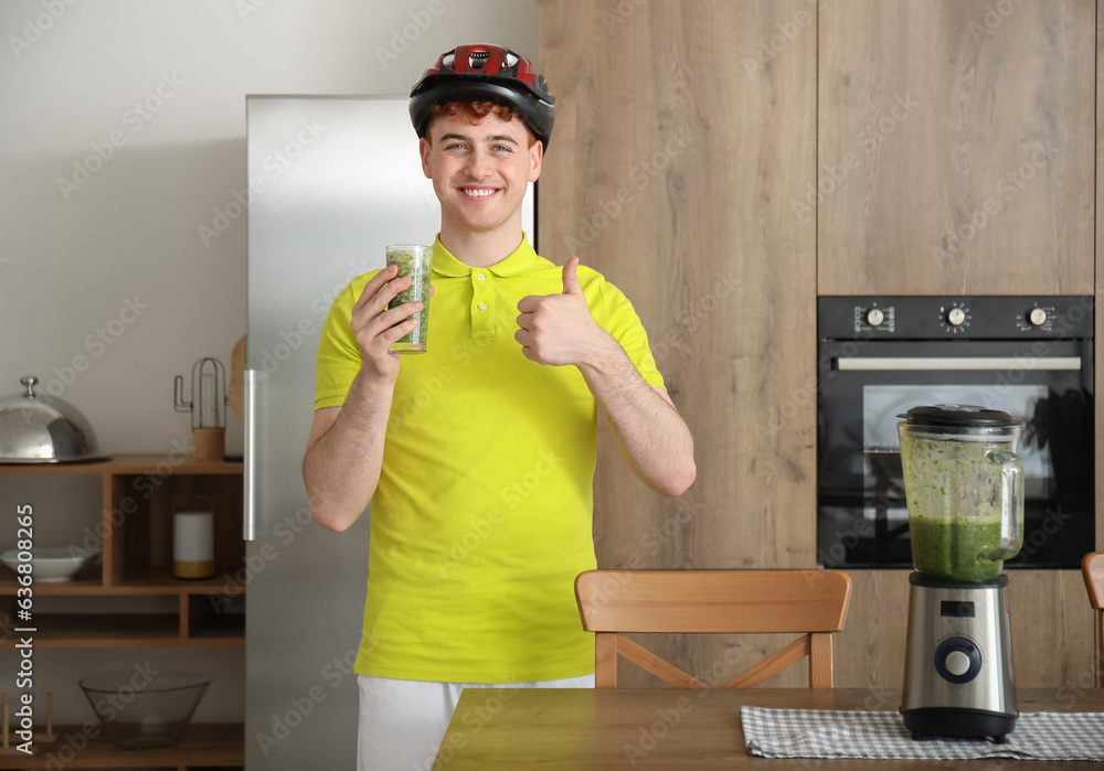 Sporty young man with glass of juice showing thumb-up in kitchen