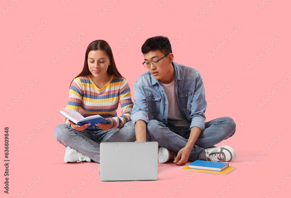 Happy students with books sitting on pink background and using laptop