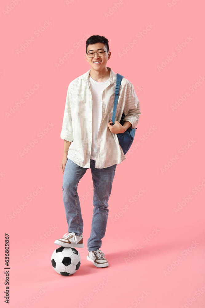 Happy male Asian student with backpack and soccer ball on pink background