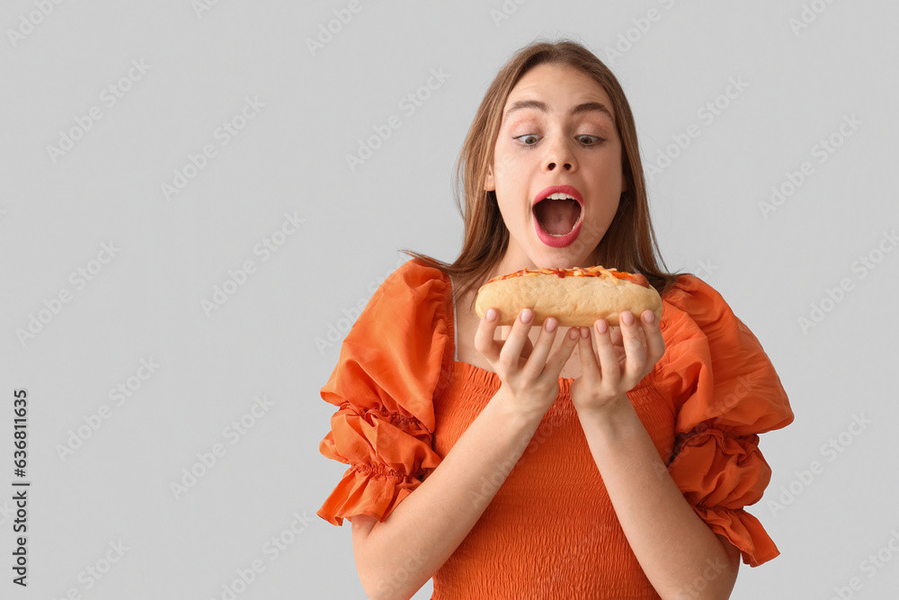 Pretty young woman eating tasty hot dog on grey background