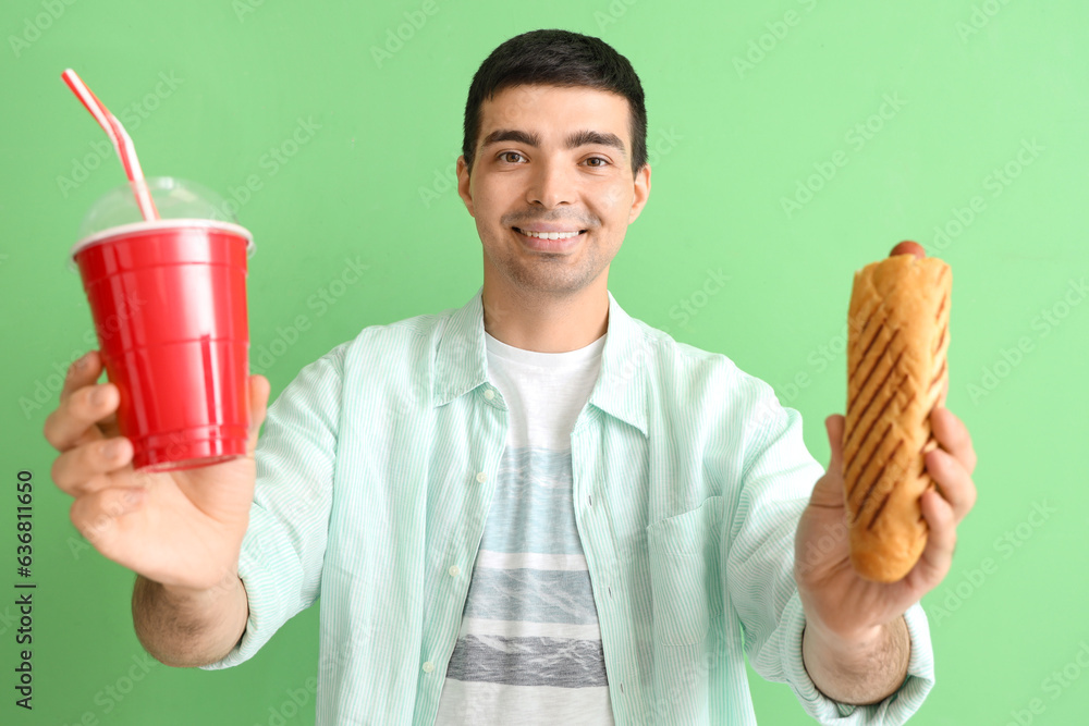 Happy young man with tasty french hot dog and drink on green background