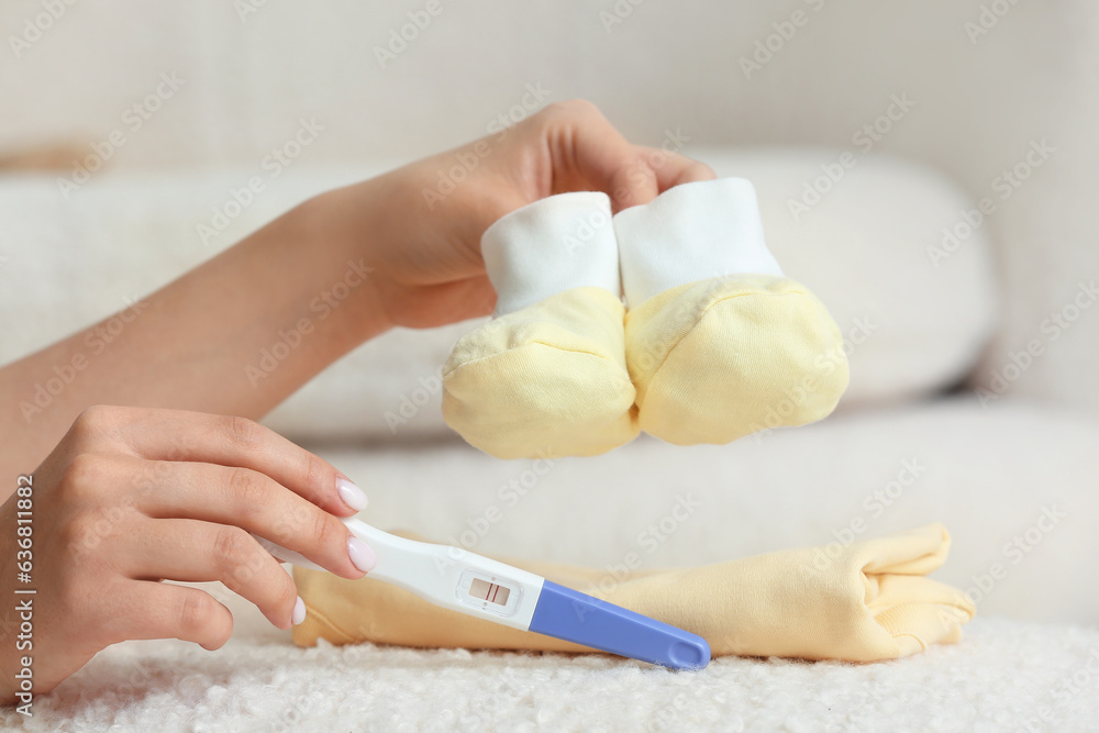 Female hands with positive pregnancy test, baby booties and clothes on sofa, closeup