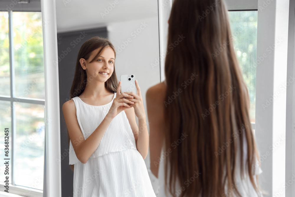 Happy little girl with mobile phone taking selfie in front of mirror at home