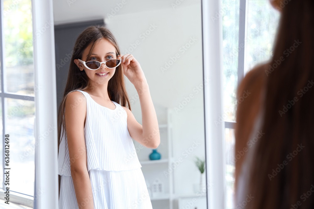 Stylish little girl with new sunglasses looking in mirror at home