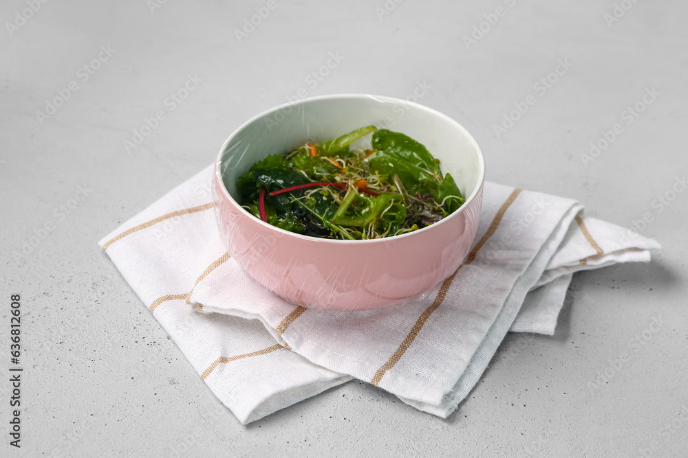 Bowl of fresh salad covered with plastic food wrap on grey background