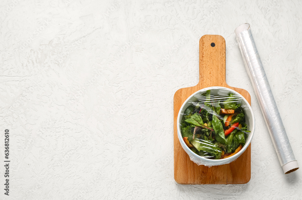 Bowl of fresh salad covered with plastic food wrap on light background