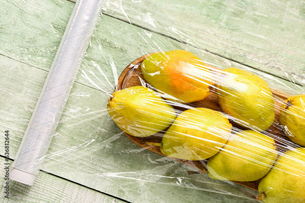 Board of fresh pears covered with plastic food wrap on green wooden background