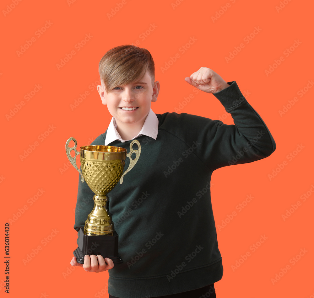 Smart little schoolboy in stylish uniform with prize cup on orange background