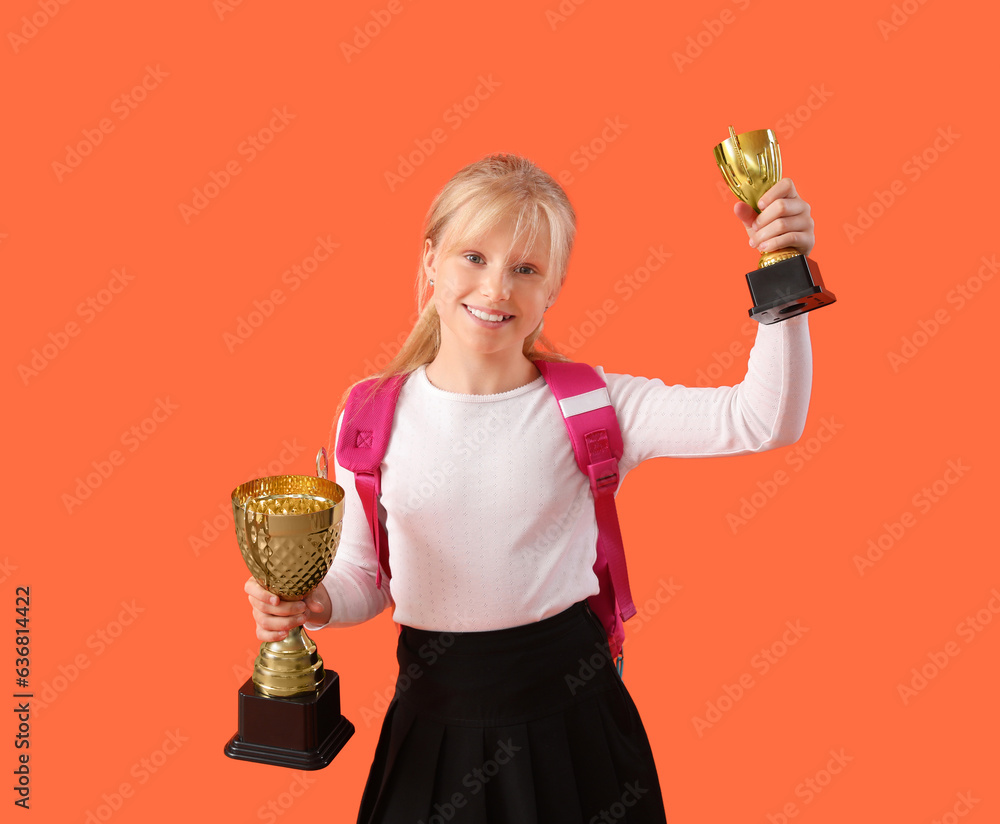 Smart little schoolgirl in stylish uniform with prize cups on orange background