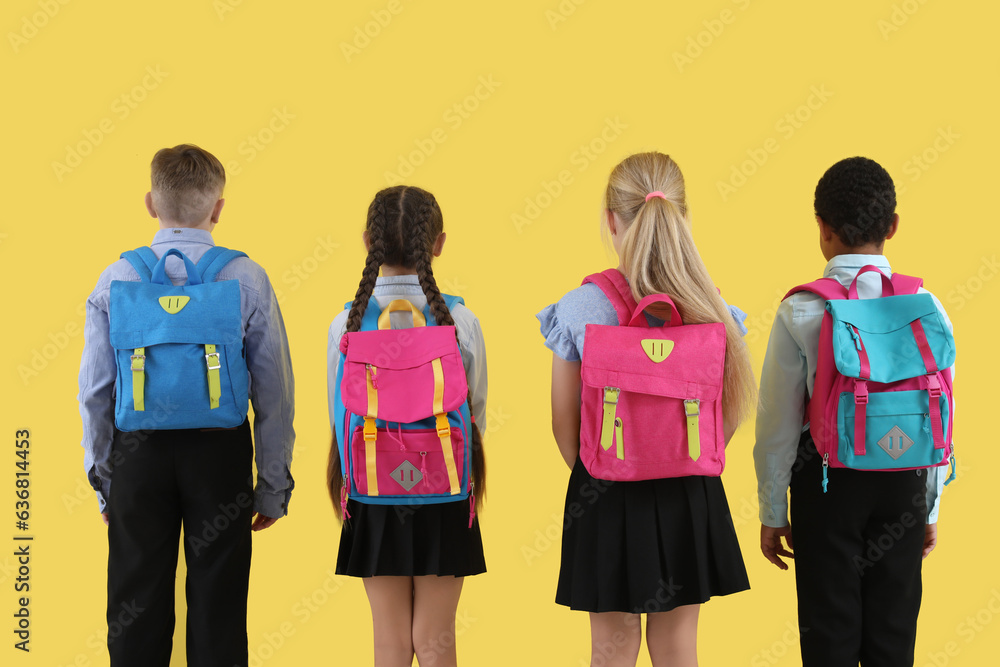 Little classmates in stylish uniform with backpacks on yellow background, back view