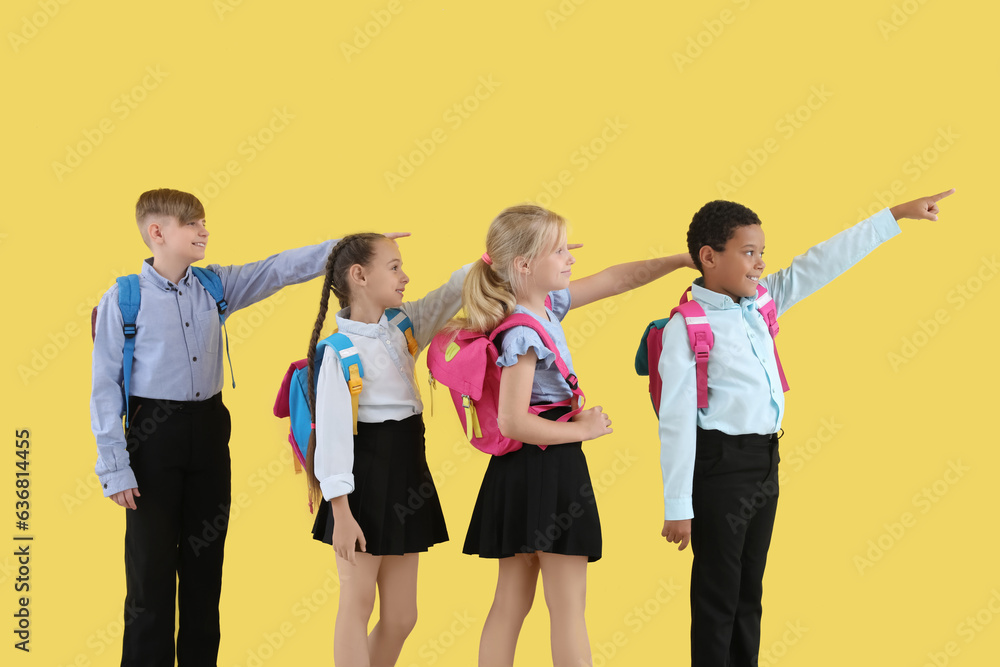 Little classmates in stylish uniform with backpacks pointing at something on yellow background