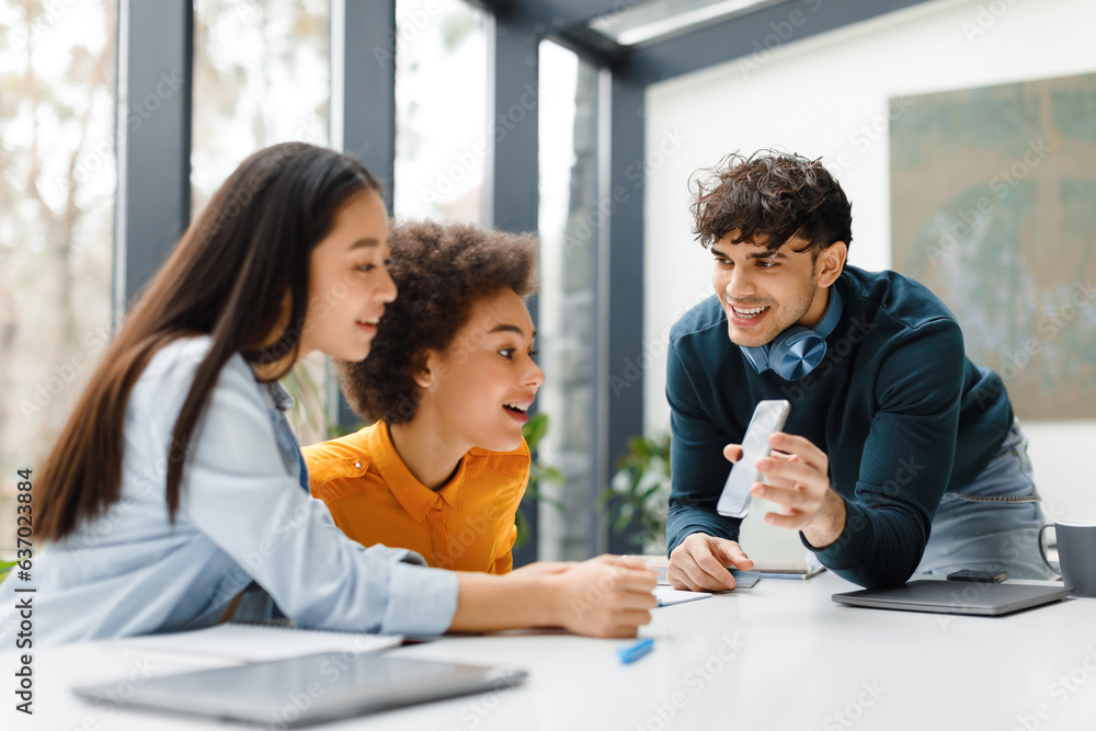 Happy european guy showing cellphone with blank screen to diverse female classmates, sitting at desk