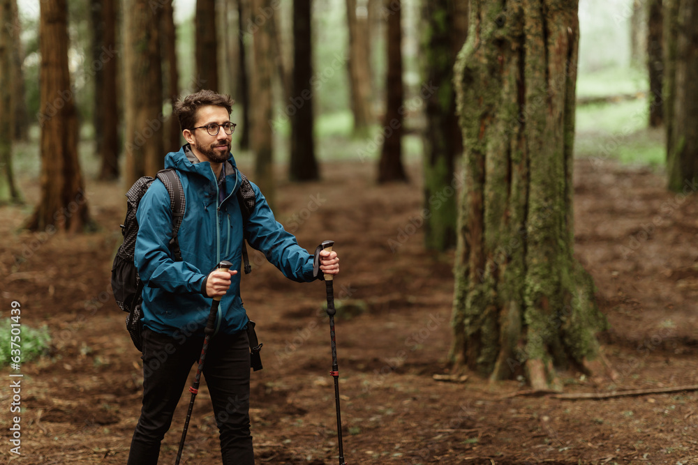 Cheerful young caucasian guy in jacket with trekking sticks walk in forest alone, enjoy trip, hiking