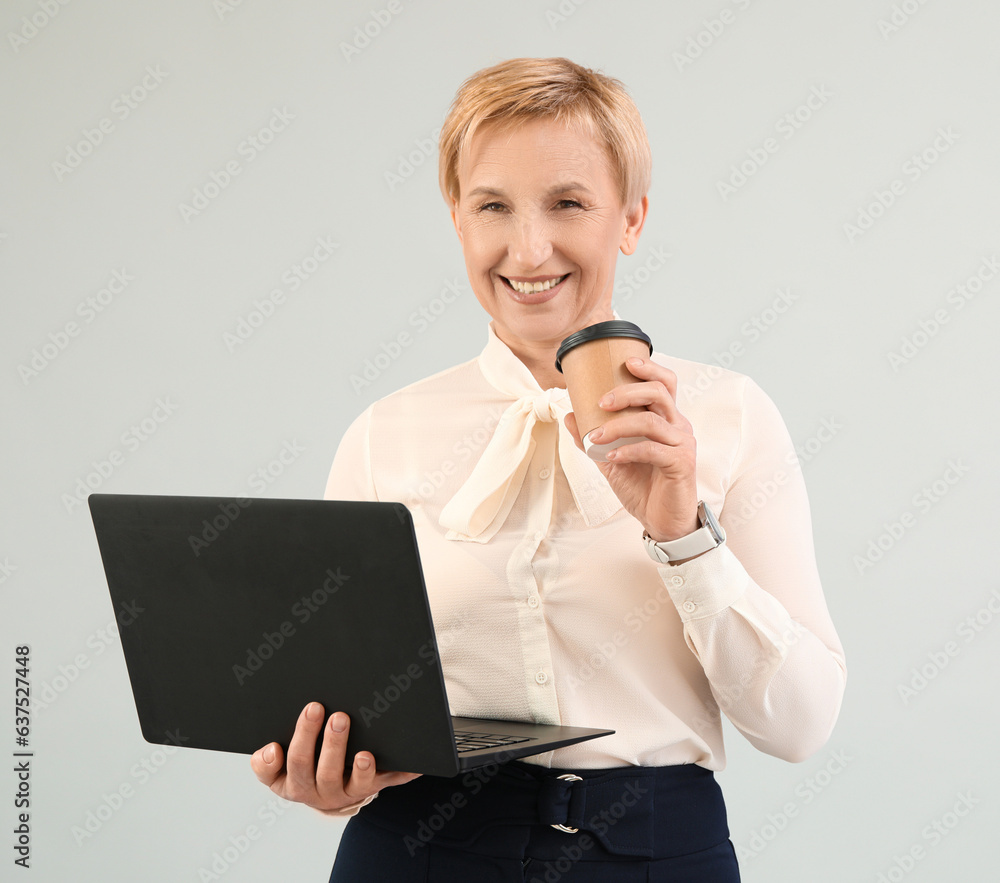 Mature businesswoman with laptop and coffee on grey background