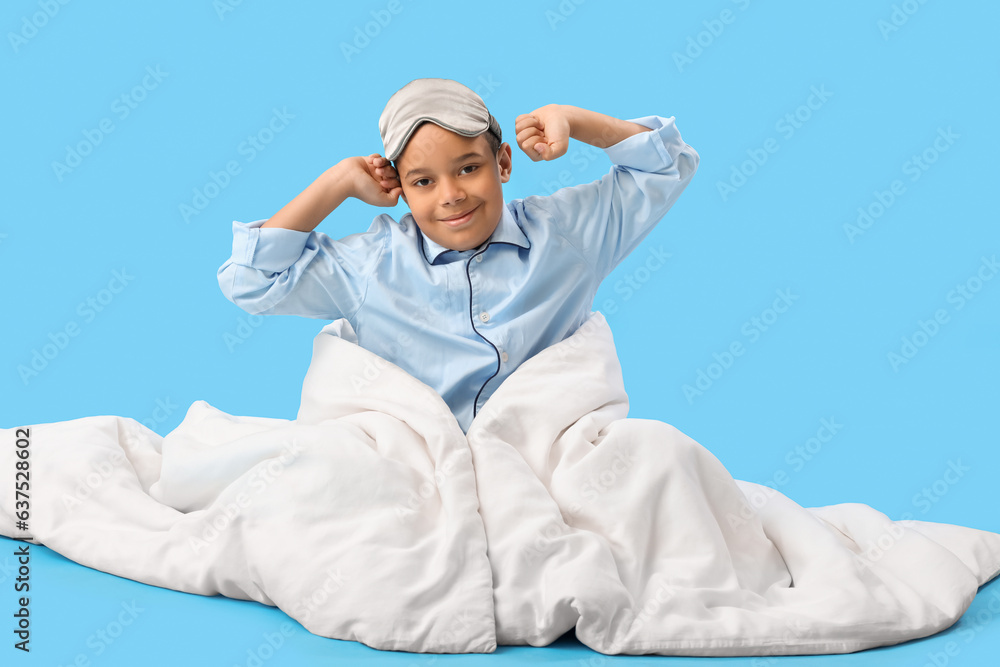 Little African-American boy with sleeping mask and soft blanket on blue background