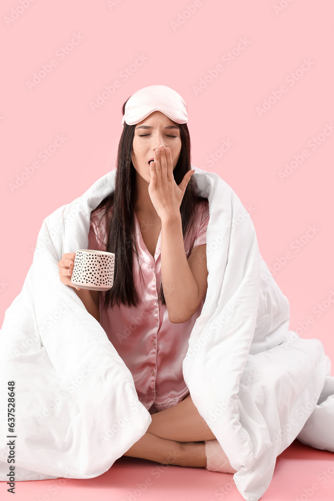 Beautiful young woman with soft blanket and cup of coffee yawning on pink background