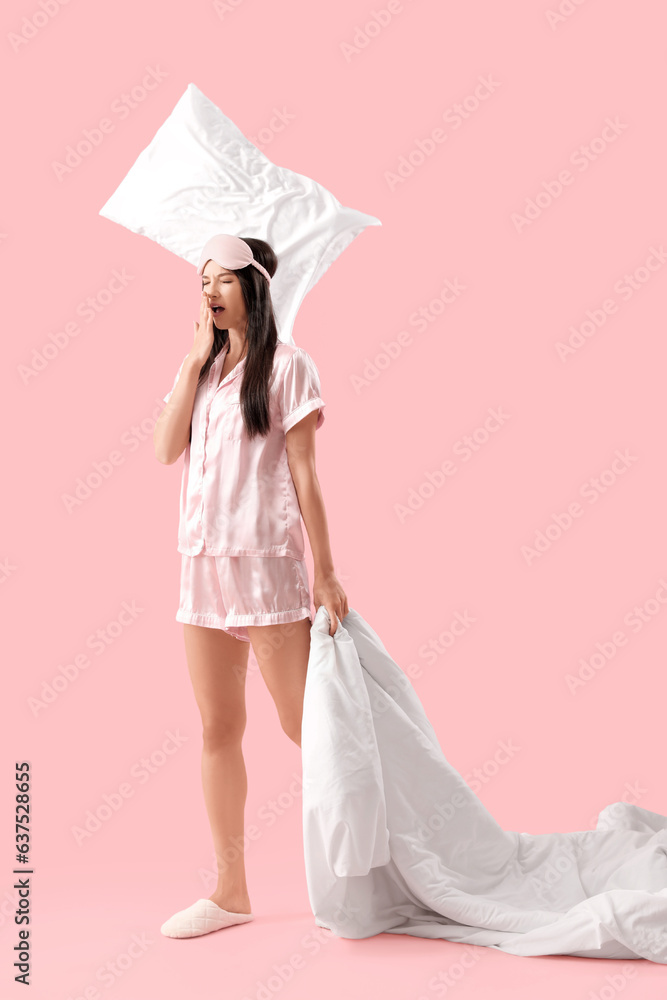 Beautiful young woman in pajamas with soft pillows and blanket yawning on pink background