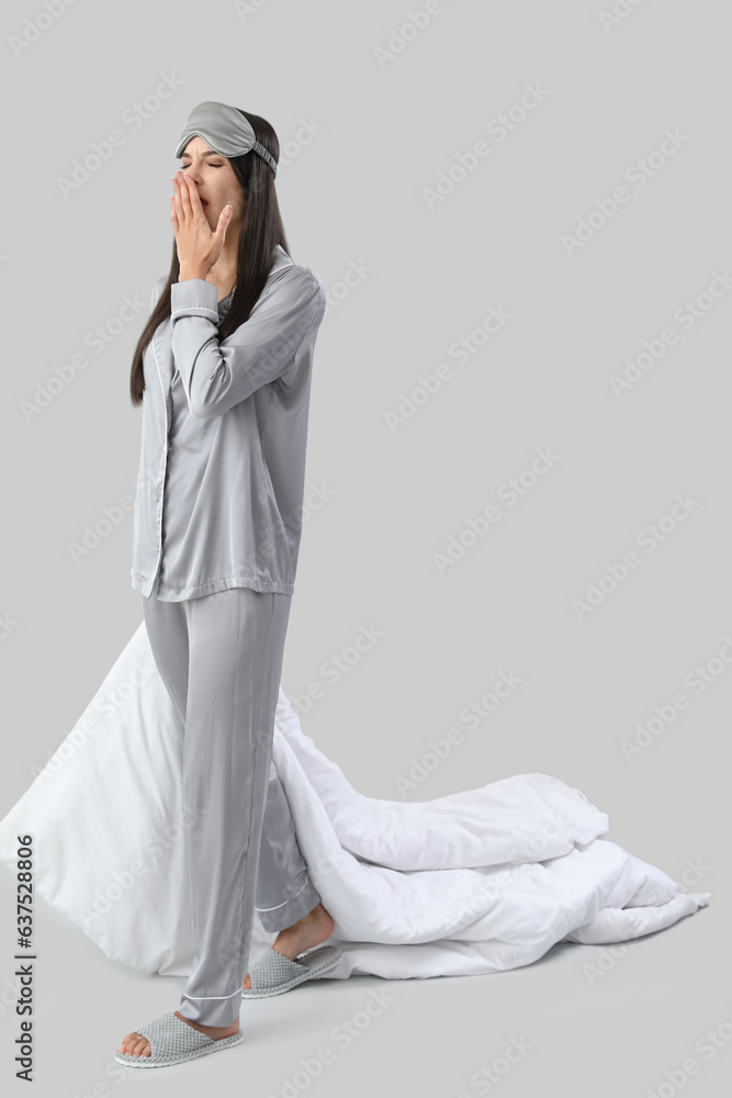 Beautiful young woman with soft blanket and pillow yawning on grey background