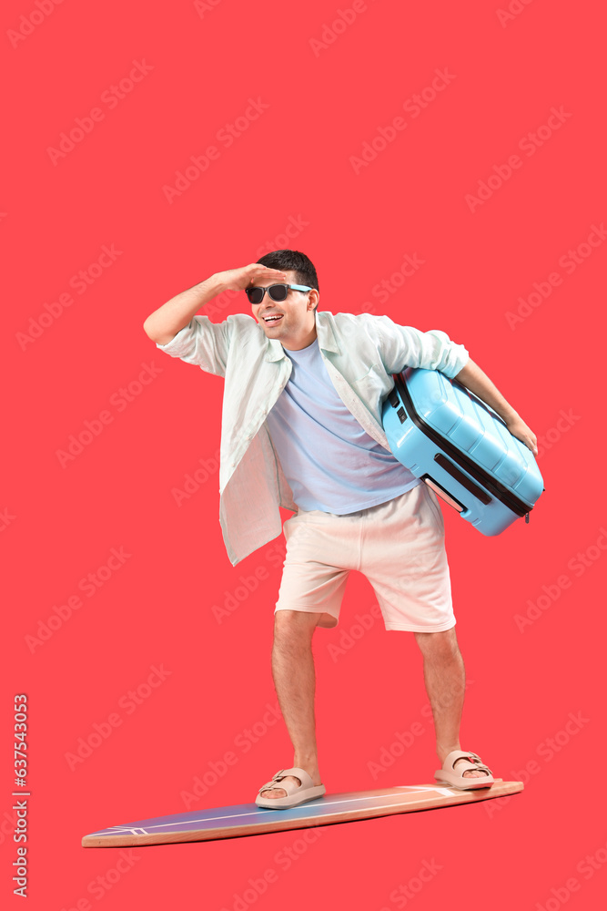 Male tourist with suitcase standing on surfboard against red background