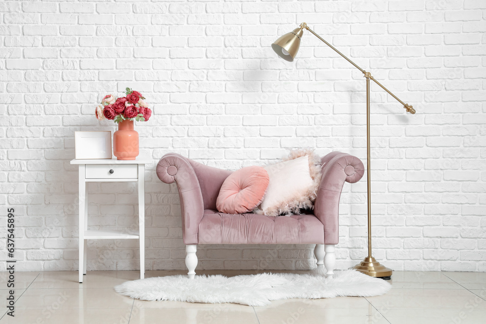 Comfortable armchair and vase with beautiful peony flowers on end table near white brick wall