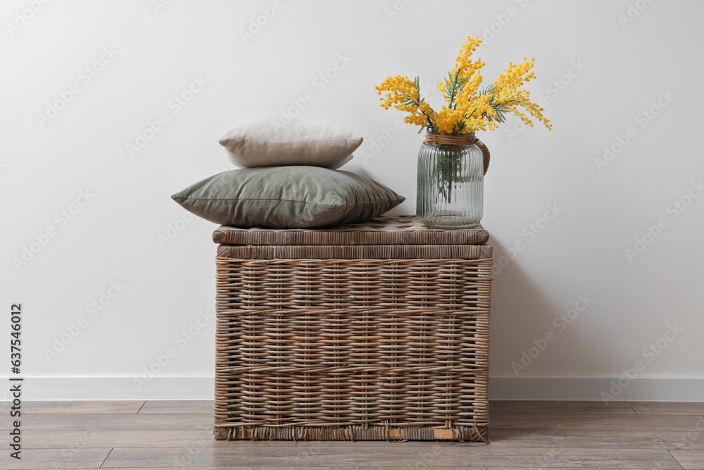 Vase with beautiful mimosa flowers and cushions on wicker basket near white wall