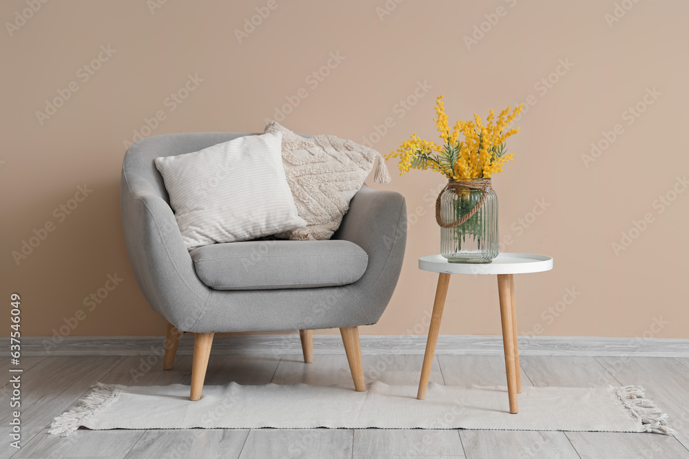 Vase with beautiful mimosa flowers on end table and grey armchair near beige wall