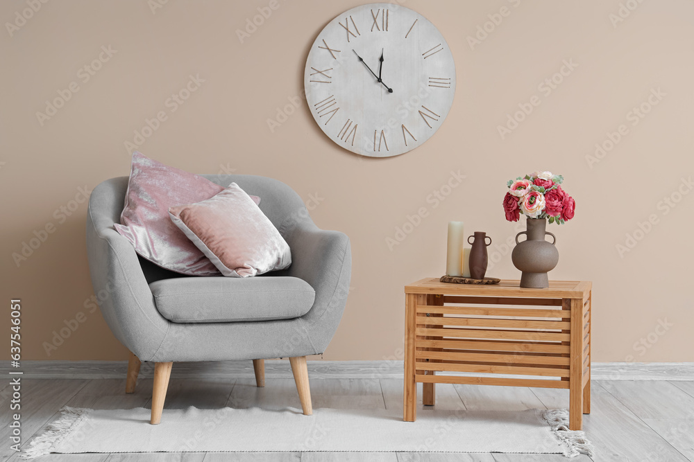 Vase with beautiful peony flowers on wooden table and grey armchair near beige wall