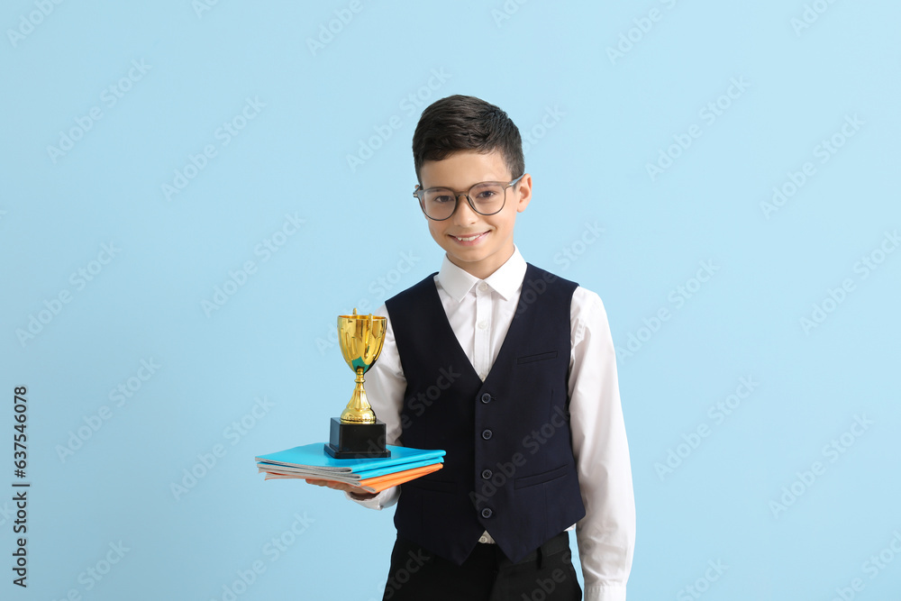 Smart little school boy with prize cup and copybooks on light blue background