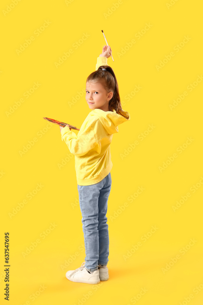 Cute little girl with paint palette and brush on yellow background