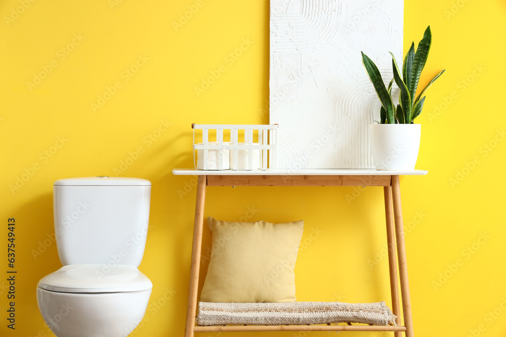 Interior of modern restroom with ceramic toilet bowl and console table near yellow wall