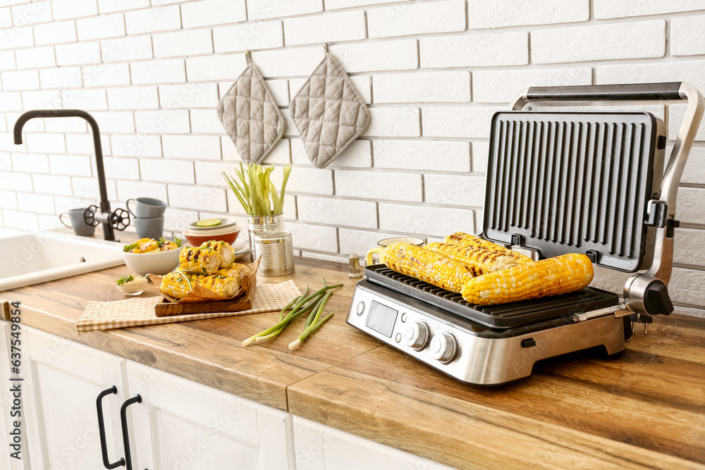 Board and electric grill with tasty corn on wooden kitchen counter