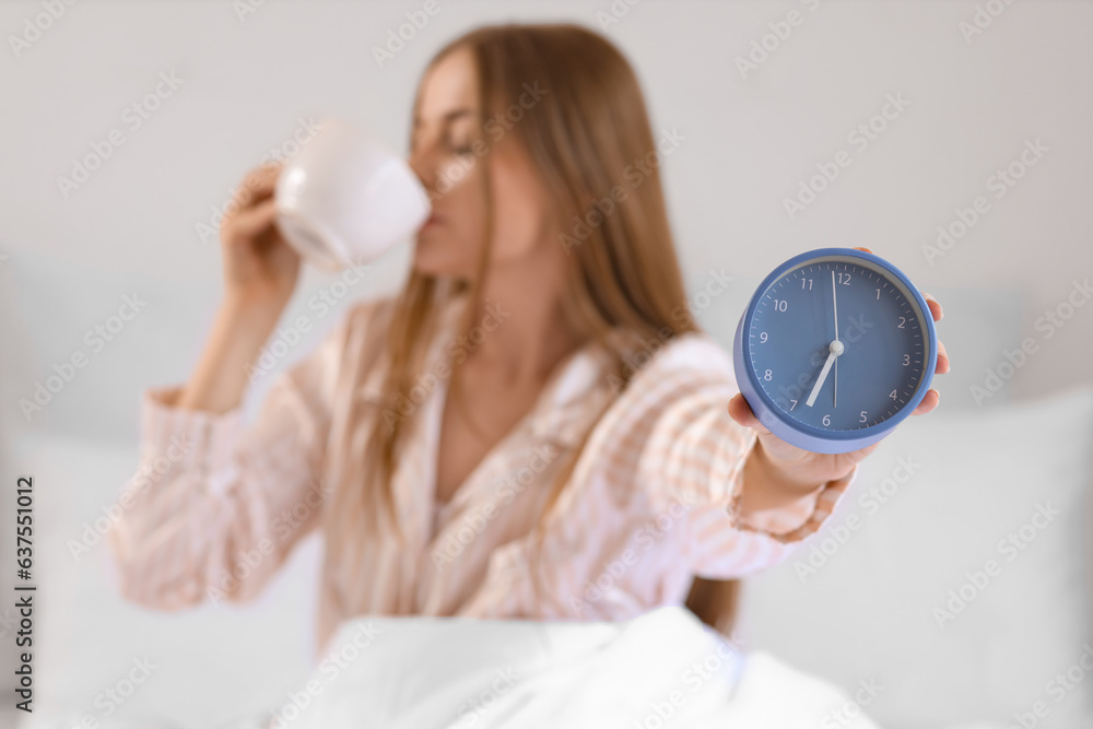 Young woman with alarm clock drinking coffee in bed at home