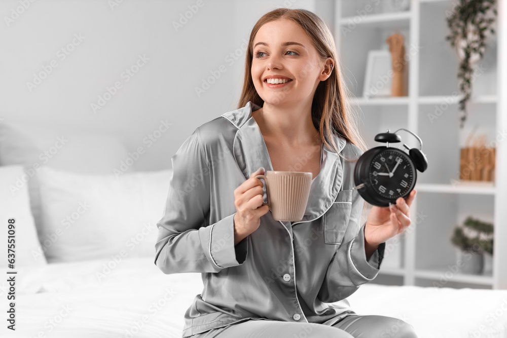 Young woman with cup of coffee and alarm clock on bed at home