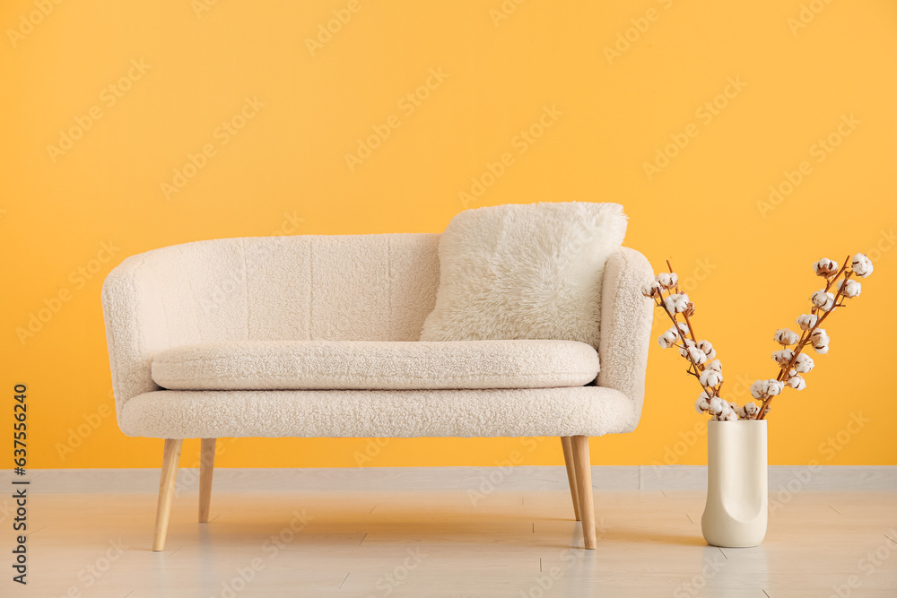 White sofa with cushion and cotton branches in vase near orange wall