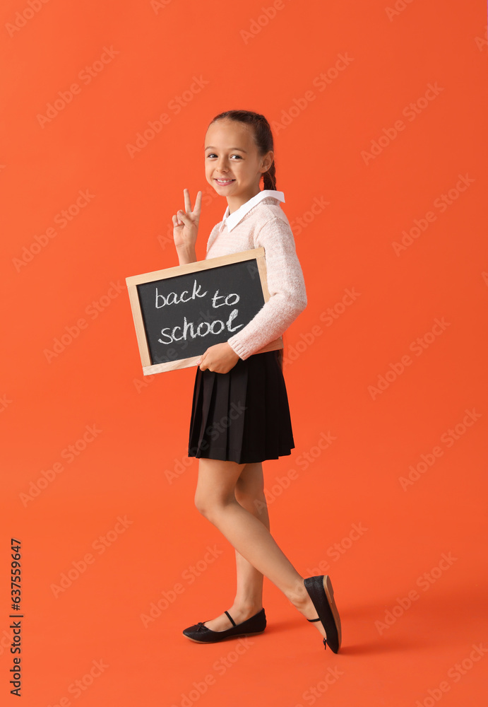 Little schoolgirl in stylish uniform holding blackboard with text BACK TO SCHOOL and showing victory