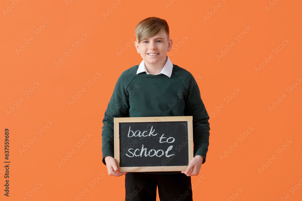 Little schoolboy in stylish uniform holding blackboard with text BACK TO SCHOOL on orange background