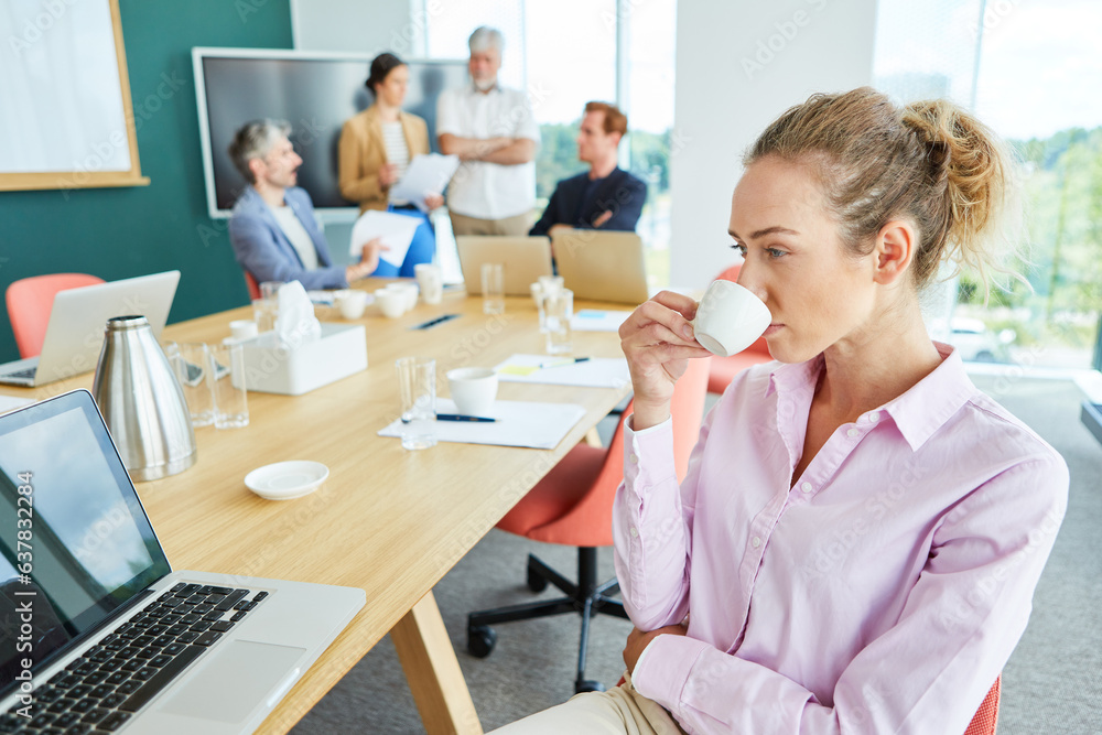 Contemplative female entrepreneur drinking coffee while staring at laptop near team discussing in co