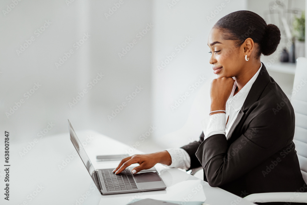 Confident black businesswoman looking at laptop and smiling, sitting at workplace in office, side vi